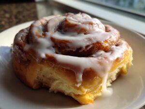 A close-up of a delicious, freshly baked protein cinnamon roll covered in thick white icing, with a bite missing from the bottom.