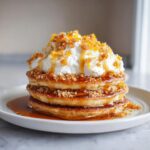 A tall stack of maple ginger pancakes topped with whipped cream, crunchy crumble, and drizzled with syrup.