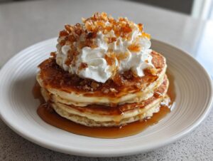 A stack of three golden maple ginger pancakes drizzled with syrup, topped with whipped cream and crunchy ginger topping.