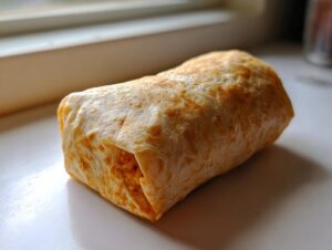 A single, tightly wrapped meal prep breakfast burrito resting on a white surface near a window.