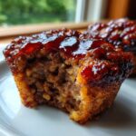 A close-up shot of a mini meatloaf muffin with a bite taken out, showing the moist interior and shiny brown glaze.