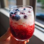 A hand holds a glass filled with a layered patriotic punch featuring red, white, and blue colors, topped with blueberries.