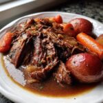 A close-up of tender, shredded slow cooker pot roast served with red potatoes and carrots in rich brown gravy.