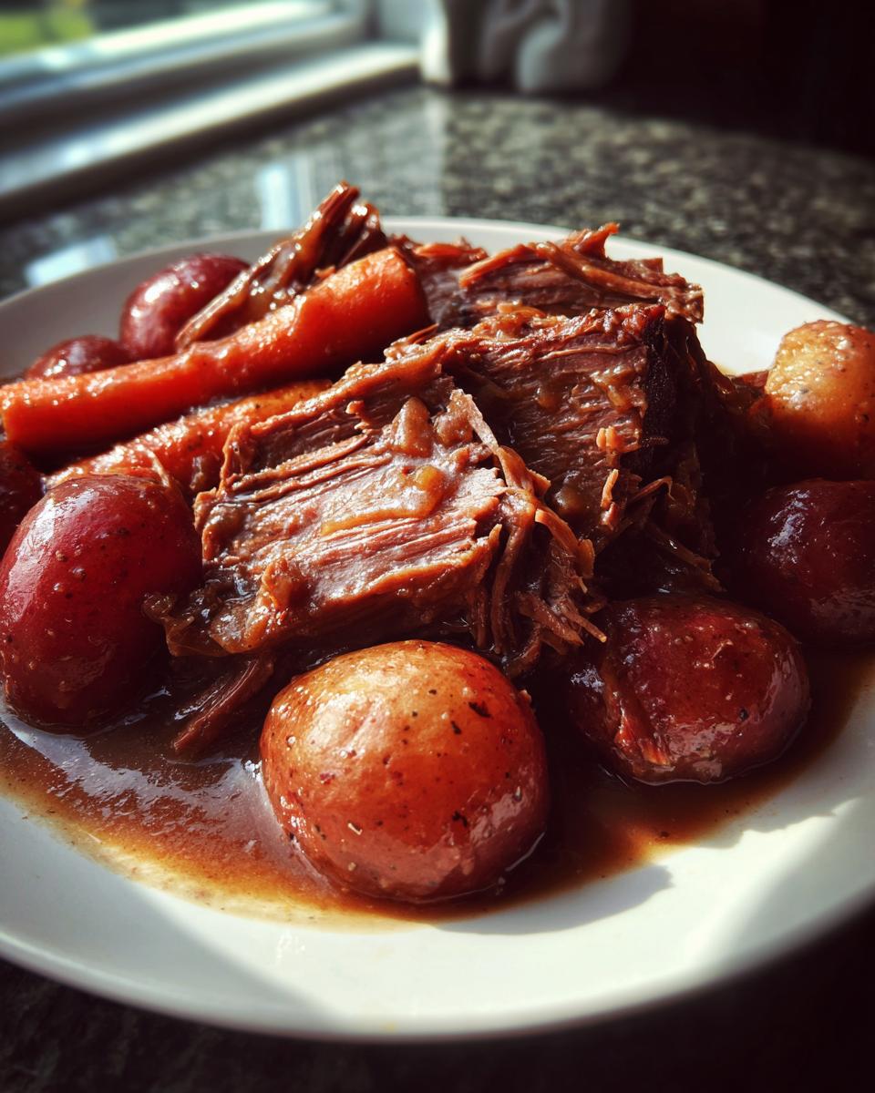 A close-up of tender, shredded slow cooker pot roast served with small red potatoes and carrots in rich gravy.