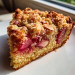 Close-up of a slice of spring rhubarb cake with almond topping, showing bright pink rhubarb filling and crumbly texture.