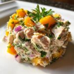 A mound of creamy tuna peaches salad mixed with diced red onion and parsley, garnished with a sprig of parsley.